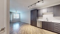 This single-floor residence showcases a thoughtfully designed open-concept layout with consistent modern finishes throughout. The kitchen serves as a central highlight, featuring dark-toned, textured cabinetry extending from floor to ceiling, white quartz-style countertops with a clean tile backsplash, and a full suite of stainless steel appliances including a refrigerator, electric range, over-the-range microwave, and dishwasher. Track lighting illuminates the kitchen and flows into the adjacent living area, which offers generous open space finished with light wood-style plank flooring and neutral gray walls. A ceiling fan provides additional comfort in the living area, which benefits from natural light through blinds-equipped windows and access to an exterior door. A secondary living or dining space also features wood-style flooring, a ceiling fan, and additional window exposure. The bedroom is carpeted and well-proportioned, with a ceiling fan and a window allowing natural light. The bathroom is finished with a dark-framed vanity mirror, matching cabinetry, white countertop, and a full tub-shower combination. Clean lines and a cohesive neutral palette create a polished, contemporary ambiance throughout the entire space.
