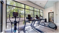 Located at Fox Carriage Dr in Raleigh, NC, this fitness center offers a thoughtfully designed, single-floor workout facility with distinct zones to accommodate a wide range of exercise routines. The space is organized into a main strength and cardio area, a cycling studio, and a functional training zone, providing a comprehensive fitness experience.

The main workout area features rubber flooring, a full-length mirror wall with a mounted flat-screen display, a power rack, free weights, benches, and a variety of cardio machines. Ceiling fans and recessed lighting keep the environment comfortable and well-lit. Large windows flood the space with natural light and offer views of the surrounding tree canopy, creating an energizing atmosphere.

A separate cycling studio is outfitted with stationary bikes and mirror-lined walls, separated from the main floor by sleek glass sliding doors. Light hardwood flooring and horizontal windows contribute to an airy, open feel.

A functional training area with hardwood flooring and a vibrant abstract floral mural serves as a creative focal point. Dumbbell racks, a medicine ball, and a ceiling-mounted air bike complete this zone. Throughout the facility, the modern aesthetic — featuring crisp white walls, dark-framed windows, and contemporary finishes — creates a clean, motivating environment suited for residents seeking a premium fitness amenity.