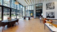 This contemporary amenity space is defined by its dramatic double-height ceilings, floor-to-ceiling glass walls, and an open, light-filled layout that creates a seamless connection to the outdoor surroundings, including views of a pool area and lush landscaping.

The main lounge area anchors the space with a full-size billiards table set atop large-format neutral floor tiles, complemented by globe pendant lights and gold-accented ceiling slats that add warmth and visual interest overhead. A striking marble feature wall serves as a refined focal point nearby.

A sophisticated sitting area showcases a bold cognac leather sofa, dark lounge chairs, and a vibrant abstract painting set against deep charcoal cabinetry with built-in shelving. Oversized windows flood this area with natural light while offering unobstructed views of the outdoor amenity deck. A ceiling fan adds a casual, comfortable touch.

Tucked alongside the main lounge is an intimate lounge nook framed by a decorative lattice partition, offering U-shaped upholstered banquette seating with leather accent pillows — ideal for relaxed gatherings.

A co-working zone, separated by glass partitions, features long communal tables with bar-height seating, a wall-mounted display screen, and an oversized ceiling fan, delivering a functional yet polished work environment. Warm wood-paneled columns and lush indoor plants complete the space, adding organic texture and life throughout.