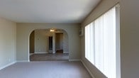 This single-floor residence showcases a thoughtfully designed interior that blends classic architectural details with practical comfort. The living area is anchored by a striking arched doorway, a timeless architectural feature that adds character and elegance while creating a natural flow between rooms. Neutral beige walls and light-toned carpeting run throughout the main living spaces, contributing to a warm and cohesive aesthetic.

Large floor-to-ceiling windows dressed with vertical blinds flood the interior with generous natural light, enhancing the sense of openness and space. A wall-mounted air conditioning unit ensures year-round climate comfort. Beyond the arch, an adjoining room continues the neutral color palette and carpeted flooring, with a ceiling fan providing additional air circulation. A glass-paneled interior door adds a subtle decorative touch, while the connected layout promotes an easy, open transition between living areas.

The overall ambiance is clean, understated, and move-in ready, offering a versatile blank canvas suited to a variety of interior styles. The consistent use of neutral finishes and classic architectural elements throughout gives the space a balanced and inviting character.
