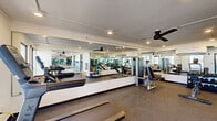 Located at 2070 McMillan Ave in North Charleston, SC, this fitness center is a well-appointed exercise facility designed to support a full range of workout routines. The single-floor space is thoughtfully laid out to maximize functionality within a compact footprint.

The exercise room is outfitted with a variety of cardio equipment, including treadmills, stationary cycling bikes with integrated display screens, and additional cardio machines. Strength training is supported by a cable/weight machine and a flat bench, while a dedicated rack of dumbbells and fitness accessories offers options for varied workouts.

Full-length wall mirrors run along multiple walls, visually expanding the space and allowing users to monitor their form during exercise. Ceiling fans are mounted overhead, contributing to air circulation and comfort. Recessed lighting provides even illumination throughout.

Durable, speckled flooring covers the space, practical for high-traffic use. Large floor-to-ceiling windows along one wall allow abundant natural light to fill the room and offer views of the surrounding outdoor area. A built-in water fountain station is conveniently positioned along an interior wall, accompanied by a wooden bench for brief rest. Storage shelving units keep accessories and supplies organized. A glass-panel entryway connects the fitness center to the broader facility.