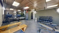 Located at 8650 Kingsbridge Dr in Saint Louis, MO, this fitness center offers a thoughtfully designed single-floor layout dedicated to health and wellness. The main exercise room is generously sized, featuring durable rubber flooring, a striking slatted wood ceiling with recessed panel lighting, and textured accent walls that lend a modern, sophisticated aesthetic. Large glass-block windows and floor-to-ceiling glass panels flood the space with natural light while providing views to the outdoors. The exercise room is well-appointed with a full suite of cardio equipment, including treadmills, an elliptical, and a stationary bike, alongside a comprehensive free weight area featuring a tiered dumbbell rack, adjustable benches, and a rack of stability balls. Full-length mirrors expand the sense of space and support proper form during workouts. Natural wood benches provide a warm contrast to the industrial finishes throughout. The two bathrooms are finished with large-format, light-toned ceramic wall tiles and mosaic tile flooring, each equipped with wall-mounted sinks, adjustable pivot mirrors, and recessed stainless steel dispensers, offering a clean and functional environment to complement the fitness facility.