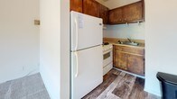Located at 8802 Three Chopt Rd in Henrico, Virginia, this single-floor unit offers a straightforward, functional layout across several distinct spaces. The living area is a notable highlight, featuring soft carpeting underfoot and a large sliding glass door that fills the room with natural light and provides direct access to a balcony. The balcony overlooks mature, leafy trees, offering a pleasant outdoor view and a sense of privacy. A wall-mounted heating unit is positioned along the base of one wall, providing climate control to the space.

The kitchen is a compact, enclosed area equipped with warm-toned wooden cabinetry, a white refrigerator, a gas range, and a sink, all set atop wood-look plank flooring that adds a durable, practical element to the space.

The bedroom is carpeted and receives natural light through a window with an outdoor view of the surrounding greenery. A louvered closet door with built-in shelving provides organized storage. A wall-mounted air conditioning unit offers additional climate control.

The bathroom features wood-look plank flooring, a tub-shower combination with white tile surround, a single vanity with an integrated sink, and standard fixtures, completing this straightforward and functional unit.
