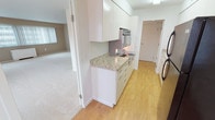 This single-floor residence offers a well-organized layout that balances functional living spaces with a clean, understated aesthetic. The galley-style kitchen is a standout feature, outfitted with crisp white cabinetry extending floor to ceiling, light-toned granite countertops, stainless steel appliances including a wall oven and electric cooktop, and a dark refrigerator. Light hardwood flooring runs throughout the kitchen, providing a warm contrast to the bright white cabinetry.

The living areas are generously sized and finished with plush neutral carpeting, giving them a comfortable, airy feel. Large windows fitted with vertical blinds allow ample natural light to fill the rooms, and baseboard heating units are positioned beneath each window. The open layout connects the living and adjacent spaces seamlessly, with clean white walls and a simple, neutral color palette throughout.

The bedroom is carpeted and includes a ceiling fan with light fixture, offering both comfort and airflow. A door from the bedroom leads directly to the bathroom, which features white subway tile walls, a glass-enclosed shower stall, and bright overhead lighting. The overall design of the space is functional and straightforward, presenting a well-maintained interior that is move-in ready.