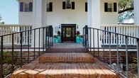 Located at 2225 Ashley River Road in Charleston, South Carolina, this single-story Southern-style building exudes classic Lowcountry charm. The exterior is defined by a crisp white facade accented with bold black shutters, tall columned porticos, and a stately entrance approached via traditional brick steps flanked by black iron railings. A rich, wood-paneled front door with glass pane inserts and sidelights serves as the focal point of the entry, offering a welcoming and distinguished first impression.

The expansive covered front porch is a standout feature, finished with elegant stone tile flooring and lined with white-railed balustrades that frame sweeping views of the lushly landscaped grounds. Mature trees draped with Spanish moss provide natural shade and reinforce the quintessential Charleston aesthetic. The porch is thoughtfully appointed with comfortable wrought-iron seating arrangements, vibrant tropical plantings in turquoise ceramic pots, and tasteful decorative accents that create an inviting outdoor gathering space.

The grounds surrounding the building are meticulously maintained, featuring colorful flower beds, manicured lawns, and brick-paved walkways that enhance the property's curb appeal. The overall ambiance blends historic Southern architecture with a warm, professionally curated environment, making this a distinctive and memorable property in the heart of Charleston.
