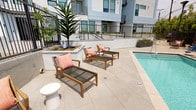Located at 8740 S La Tijera Blvd in Los Angeles, CA 90045, this outdoor amenity level at Eastway offers a thoughtfully designed communal space that blends indoor comfort with open-air living. The expansive tiled terrace serves as the central hub, featuring large-format light stone pavers that extend throughout and unify the various activity zones.

At the heart of the space is a resort-style pool deck complete with teak chaise lounges accented with coral cushions, ideal for relaxing in the Southern California sun. Adjacent to the pool, a dedicated BBQ and outdoor kitchen station provides a built-in grill and prep area, alongside a dining zone furnished with a wood table and vibrant orange chairs set atop a decorative rug.

A standout architectural feature is the oversized glass bi-fold door system that opens the indoor lounge directly onto the terrace, creating a seamless indoor-outdoor flow. The interior lounge is furnished with contemporary navy and wood seating, a wall-mounted TV, and a bar area, making it a versatile social space.

Lush landscaping is woven throughout, with raised planter beds showcasing tropical palms, bird-of-paradise, philodendrons, and ornamental trees that add vibrant greenery and a resort-like ambiance. The surrounding multi-story white and blue building frames the space, contributing to a modern, cohesive aesthetic.