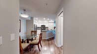 This single-floor residence offers a thoughtfully designed open-concept layout, connecting the kitchen, dining, and living areas into one cohesive space. Light wood-tone flooring runs throughout the main living areas, adding warmth and visual continuity.

The kitchen is outfitted with white shaker-style cabinetry, a gray tile backsplash, dark countertops, and a full suite of stainless steel appliances, including a range with microwave above and a refrigerator. A curved peninsula with an undermount sink provides additional workspace and serves as a natural divider between the kitchen and the adjoining dining area, where a glass-top table and cushioned chairs are comfortably situated beneath a pendant light.

The living room features a ceiling fan, hardwood-style flooring, and an open flow to both the kitchen and hallway. A dedicated laundry area includes a full-size washer and dryer with overhead shelving and a window for natural light.

The bedroom is carpeted and spacious, featuring a ceiling fan, soft neutral walls, and access to a walk-in closet with wire shelving. The bathroom features wood-look flooring, white cabinetry, a dark countertop, and a shower with curtain enclosure.