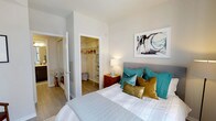 This single-floor space showcases a thoughtfully designed bedroom that combines functionality with contemporary style. The room is finished with warm wood-look flooring that runs seamlessly throughout, complementing the crisp white walls and creating a bright, clean atmosphere. A generously sized bed serves as the focal point, dressed in neutral linens and accented with teal and gold throw pillows that add a pop of color against the understated backdrop. A framed piece of abstract artwork adorns the wall above the headboard, contributing to the room's modern character. A warm-toned wooden nightstand with a table lamp provides practical bedside storage and soft ambient lighting. One of the room's standout features is the walk-in closet, accessible directly from the bedroom. It is equipped with built-in shelving and hanging rods, offering ample organized storage space. An additional door visible from the room leads to what appears to be a mirrored space, likely a bathroom or hallway, further enhancing the sense of connectivity within the layout. Overall, the space balances comfort, storage, and modern design in a cohesive and inviting manner.