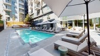 Located at 1120 W 6th St in the heart of Downtown Los Angeles, this outdoor amenity space delivers a resort-style experience within an urban setting. The centerpiece is a sparkling rectangular pool with crystal-clear blue water, surrounded by an expansive pool deck finished in smooth concrete. A generous arrangement of cushioned lounge chairs and chaise lounges lines both sides of the pool, offering ample space for relaxation under the Southern California sun. Large white umbrellas are positioned throughout the deck, providing shade and a clean, modern aesthetic. A standout feature of the space is the collection of freestanding wooden cabanas, constructed with warm-toned slatted timber panels and open on one side to allow airflow. Each cabana is outfitted with plush seating and contemporary accent tables, creating semi-private retreats ideal for lounging or socializing. A lush living green wall runs along the perimeter, adding a vibrant natural element that contrasts beautifully with the modern architecture of the surrounding multi-story building. The building's facade, visible from the pool deck, features a striking mix of dark and light exterior finishes with private balconies above. An adjacent indoor lounge area with glass walls connects seamlessly to the outdoor deck, enhancing the overall flow of the space.