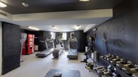 This dedicated fitness center is designed with both functionality and motivation in mind. The space is defined by its bold, dark chalkboard-painted walls adorned with handwritten motivational phrases such as "Don't Quit," "Build Your Legacy," and "Time to Workout," creating an energizing and inspiring atmosphere throughout.

The cardio zone is well-stocked with multiple treadmills, a Precor elliptical trainer, and a stationary bike, all positioned to take advantage of available natural light from windows along one wall. Overhead fluorescent lighting ensures the entire space is brightly and evenly lit.

The strength training area features a full rack of dumbbells in varying weights, a adjustable workout bench, medicine balls, and a ball rack, offering a comprehensive range of free weight and functional fitness options. A large wall-mounted mirror spans one section of the room, allowing users to monitor their form during exercises.

Practical amenities include a Coca-Cola vending machine for post-workout refreshment and convenient access via a glass-panel entry door. The floor is finished in a durable, light-toned surface suited for high-traffic fitness use. The overall design balances an industrial aesthetic with a motivating, community-oriented atmosphere.