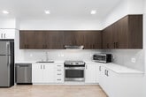 This contemporary single-floor unit is designed with clean lines and a modern aesthetic throughout. The open-concept kitchen and living area serves as the heart of the space, featuring light wood-tone flooring that extends seamlessly across the room. The kitchen is well-appointed with a two-tone cabinet design — crisp white lower cabinets paired with warm dark wood upper cabinets — complemented by a white tile backsplash and light countertops. Stainless steel appliances, including a refrigerator, dishwasher, and range with hood, provide a polished, functional finish. Recessed lighting illuminates the space from above, while a large floor-to-ceiling window at the end of the living area floods the room with natural light and offers views of the surrounding urban environment. A separate bedroom features soft neutral carpet, a tall vertical window that invites additional natural light, and clean white walls that create a calm, restful atmosphere. The bathroom is finished in a minimalist style, with wood-look flooring, white walls, a combined tub and shower enclosure with subway-style tile surround, and a grab bar for added accessibility. Throughout the unit, the consistent use of light tones and modern finishes creates a cohesive, contemporary living environment.