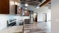 Located at 1600 O St in Lincoln, Nebraska, this 2-bedroom, 2-bathroom loft-style residence offers a compelling blend of industrial character and modern finishes across a single floor. The space is defined by its striking exposed wood ceiling joists and brick, which lend a raw, historic authenticity throughout the main living areas and primary bedroom. Large, multi-pane windows flood the interior with natural light and frame views of the surrounding neighborhood's mature trees.

The primary bedroom is generously proportioned, featuring neutral carpet flooring, a ceiling fan, and the same exposed overhead structure that gives the space its distinctive industrial aesthetic. A dedicated corridor connects to one of the two full bathrooms, which is equipped with a walk-in shower featuring built-in shelving, a sliding glass door, and a linear drain. Open shelving within the bathroom provides practical storage. Dark-toned wood cabinetry and granite-style countertops add a refined contrast to the otherwise clean, white palette.

The second bedroom offers a quieter, more enclosed feel with plush carpeting, tall ceilings, and ample natural light through its own set of large windows. Sleek, dark-framed glass doors and hardwood-style luxury vinyl plank flooring in the common areas tie the overall design together, creating a cohesive urban loft aesthetic throughout.