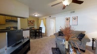 This single-floor, 1-bedroom, 1-bathroom unit offers a well-organized layout designed to maximize functionality. The open-concept living and dining area serves as the heart of the space, seamlessly connecting the two zones for effortless everyday living. The living area features plush wall-to-wall carpeting, a ceiling fan with a light fixture for comfort and illumination, and ample space for standard furniture arrangements. A glass-top coffee table and media console complement the relaxed, casual ambiance. Adjacent to the living space, the dining area accommodates a full dining set beneath warm, yellow-toned walls accented with colorful artwork, adding a welcoming pop of color. A window dressed with golden curtains allows natural light to filter into this area, brightening the room. The entry door is conveniently positioned between the living and dining zones, offering a practical flow throughout the unit. The overall aesthetic is warm and approachable, with neutral tones in the main living space complemented by accent colors in the dining area, creating a balanced and inviting atmosphere throughout.