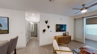 This single-story residence showcases a contemporary design with clean lines, white walls, and light wood-look flooring throughout the main living areas. The open-concept living room is a welcoming space, featuring a ceiling fan, a wall-mounted television, decorative geometric wall planters, and a sliding barn door that leads to an adjacent area filled with natural light from large windows.

The kitchen is thoughtfully designed with granite countertops, two-toned cabinetry pairing white upper cabinets with sage-green lower cabinets, and stainless steel appliances. A peninsula with pendant lighting and bar seating creates an ideal space for casual dining and entertaining.

The bedroom is a serene retreat with plush carpet flooring, large windows dressed with elegant draping curtains, a ceiling fan, and ample natural light. The en-suite bathroom continues the granite countertop theme, featuring grey cabinetry, a shower-tub combination, and direct access to a spacious walk-in closet with built-in shelving and hanging storage. Recessed lighting throughout the unit adds a polished, modern touch to the overall ambiance.