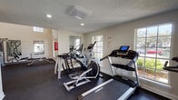This fitness center is thoughtfully designed across two connected zones, offering a comprehensive workout experience for users of all fitness levels. The cardio room features a textured ceiling with recessed lighting and is lined with large, multi-pane windows that flood the space with natural light and provide views of the surrounding greenery. It is equipped with Spirit-brand treadmills, elliptical cross-trainers, and a stationary bike, all positioned on dark rubber flooring. A full-length mirror on one wall enhances the sense of space and allows for form-checking during workouts. The adjoining strength training room boasts impressively high ceilings with tall windows that draw in additional natural light. This area is outfitted with a multi-station cable weight machine, a Hoist adjustable weight bench, a rack of dumbbells in varying sizes, and a body weight scale. Wall-mounted mirrors assist with technique and contribute to the open, airy feel of the room. The dark rubber flooring throughout both areas provides traction and durability. Together, the two rooms create a well-rounded fitness facility that balances cardio and strength training in a bright, functional environment.