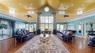 Located at 115 112th Ave N in Saint Petersburg, Florida, this single-floor community space makes a strong architectural impression from the moment you enter. The centerpiece is a grand living room boasting soaring vaulted ceilings finished in a warm golden yellow, accented by teal walls that lend a distinctly coastal Florida character. A striking circular clerestory window crowns the peak of the vaulted ceiling, while rows of tall windows and French doors flood the interior with abundant natural light and frame lush outdoor greenery. Dual ceiling fans provide comfort throughout the expansive room, which features rich hardwood flooring and is anchored by an elegant, large-format area rug. Comfortable leather seating arrangements and decorative tropical plants reinforce the welcoming, resort-like ambiance. Floral drapery panels add a playful, colorful touch that complements the overall coastal aesthetic. The dedicated office/conference room offers a more focused environment, furnished with a substantial hardwood conference table and upholstered rolling chairs, with plantation-shuttered windows, teal walls, and granite-topped built-in cabinetry providing both functionality and style. The entrance and connecting hallway complete this well-appointed space, offering a smooth and polished transition between areas.