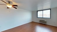 Located at Namataur Towers, this single-floor space offers a straightforward and functional layout with a clean, minimalist aesthetic. The main living area is characterized by smooth white walls and warm hardwood-style flooring that extends throughout, lending a cohesive and inviting feel to the space. A ceiling fan is mounted overhead, providing efficient air circulation throughout the room. Natural light enters through a window fitted with horizontal blinds, offering views of the surrounding urban environment. A wall-mounted air conditioning unit sits beneath the window, ensuring year-round climate control. The white ceiling and walls create a bright, neutral backdrop that makes the space feel open and adaptable to a variety of furnishing styles. The overall layout is simple and efficient, making practical use of the available floor area. The space is well-suited for those seeking a comfortable, low-maintenance living environment within the Namataur Towers building.