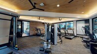 This contemporary fitness center offers a thoughtfully designed workout environment that balances both aesthetic appeal and functional utility. The space features a distinctive tray ceiling clad in warm wood paneling, accented by soft ambient lighting that runs along the ceiling's perimeter, creating an inviting atmosphere throughout. Large black-framed glass partitions and windows allow natural light to filter into the space while providing views of the surrounding exterior areas, contributing to an open, airy feel.

The fitness center is organized into distinct workout zones. The cardio area is outfitted with multiple treadmills and an elliptical trainer, positioned along the perimeter to maximize floor space. A multi-station strength machine serves as a central fixture, offering a range of resistance training options. A dedicated free weights zone features power racks, a flat bench, and barbell sets, all positioned on protective rubber flooring for safe and comfortable lifting sessions.

Durable, textured carpet tiles cover the main floor, providing comfort and traction underfoot. Oversized ceiling fans ensure consistent airflow throughout the space, enhancing comfort during workouts. Framed artwork and carefully considered design details add a polished, upscale character to the facility, making it a well-rounded destination for a comprehensive fitness routine.