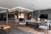 Located at Westminster Way N in Shoreline, Washington, this contemporary single-floor amenity space is thoughtfully designed to accommodate a variety of social and recreational activities. The open-concept layout flows seamlessly across wood-look tile flooring, connecting distinct functional zones while maintaining a cohesive, modern aesthetic throughout.

The kitchen area serves as a central hub, featuring a generous island with bar seating, warm wood-paneled ceilings with pendant lighting, dark cabinetry, stainless steel appliances, and a striking textured tile backsplash. Adjacent to the kitchen, a spacious lounge area offers comfortable sectional seating, a large wall-mounted television, and direct access to an outdoor terrace through expansive floor-to-ceiling glass doors and windows.

A dedicated game and recreation zone includes a pool table and high-top seating, accented by bold planter dividers that add greenery and visual interest. A flexible co-working and dining area provides multiple seating configurations with high-top tables, benches, and task chairs, all bathed in natural light from large perimeter windows framing lush Pacific Northwest views. The overall ambiance is sleek yet inviting, blending contemporary design with functional versatility.