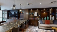Located at 4200 N Carlin Springs Rd in Arlington, VA, this thoughtfully designed amenity lounge offers a sophisticated and multi-functional gathering space across a single floor. The space is anchored by striking oxidized cylindrical columns that lend an industrial-chic character throughout. Wide-plank hardwood flooring flows through the open layout, complementing the warm tones of the wood-paneled walls and built-in shelving.

A full billiards table takes center stage in the main area, while a dark granite-topped bar with leather upholstered barstools serves as a social hub, illuminated by elegant pendant lighting with sculptural designs. Adjacent to the bar, a lounge nook features built-in bookshelves lined with curated volumes and decorative objects, paired with plush seating and an artfully patterned area rug.

Vibrant rust-orange lounge chairs clustered around a glass-top table create an inviting conversation area near a glass-railed mezzanine overlooking a double-height atrium. The atrium showcases a dramatic cascading globe chandelier framed by floor-to-ceiling windows that flood the space with natural light and exterior views. A wood-paneled elevator bay adds warmth and texture, while curated artwork and sculptural accents throughout reinforce the space's refined, contemporary aesthetic.