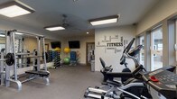 This fitness center is a comprehensively outfitted single-floor space designed to support a full range of workout routines. The room is finished with durable rubber-backed flooring and illuminated by large flush-mounted fluorescent panels, supplemented by a ceiling fan that helps maintain a comfortable training environment. Natural light enters through multiple windows, offering pleasant views of an outdoor pool, patio seating, and landscaped grounds beyond a fenced perimeter.

The space is divided into distinct functional zones. A strength training area features a Smith machine with weight plates, a flat workout bench, and a well-stocked dumbbell rack with hex weights in varied sizes. Full-length mirrors line one wall, enhancing the sense of space and allowing users to monitor their form. A cardio zone is equipped with elliptical trainers, a recumbent bike, and a treadmill. Additional equipment includes medicine balls, stability balls, and resistance accessories stored on a dedicated rack.

A bold fitness-themed typographic mural anchors one wall, adding energy and motivation to the space. Colorful artwork provides visual contrast on another wall. A touchless waste bin, towel hooks, and posted guidelines reflect thoughtful amenity management. A glass-panel door connects to an adjacent room, likely restroom or locker facilities.