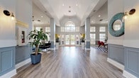 This expansive single-floor community clubhouse is defined by its dramatic vaulted ceilings and an open, airy floor plan that encourages social interaction throughout the space. The grand entrance foyer sets the tone immediately, with a signature arched transom window above French doors that frame views of an outdoor pool area beyond. Tall, grid-pane windows line the walls throughout, flooding every corner with abundant natural light and reinforcing the sense of openness.

Wide-plank hardwood-style flooring runs continuously across the entire space, visually unifying the distinct functional zones. Structural columns with wainscoted bases add architectural definition without interrupting the flow between areas. The central lounge zone features multiple seating arrangements, including plush navy sofas, modern accent chairs in charcoal and teal, and bold patterned area rugs in deep blue tones, all complemented by colorful abstract artwork displayed on the walls.

Adjacent to the lounge, a dedicated dining area offers a warm wood table surrounded by vivid red chairs, set atop a striking geometric black-and-white rug, with a designer open shelving unit adding character. Lush tropical plants in tall white planters are positioned throughout, providing natural accents. Ceiling fans are installed across the space, enhancing comfort while adding to the relaxed yet sophisticated ambiance of this well-appointed gathering facility.