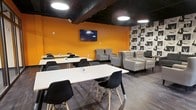 This single-floor space makes a strong visual impression through its bold design choices and versatile layout. The most striking feature is the vibrant orange accent wall, which anchors the room and creates an energetic atmosphere balanced by a dark, exposed ceiling that adds an industrial edge. Circular flush-mounted ceiling lights provide even, modern illumination throughout the space.

The room is divided into two distinct seating zones. A casual lounge area along one side features light grey upholstered armchairs and a tufted sofa arranged around a low table, offering a relaxed setting for conversation or informal meetings. A graphic feature wall with a repeating black-and-white pattern adds visual interest to this corner of the room.

The dining or collaborative zone consists of multiple long white tables paired with contemporary black molded chairs on natural wood legs, arranged to accommodate groups of varying sizes. A wall-mounted flat-screen television is positioned prominently above the orange wall, making the space suitable for presentations or group viewing.

Wood-look flooring runs throughout, tying the two zones together and contributing warmth to the otherwise bold, urban aesthetic. Overall, the space feels dynamic, functional, and well-suited for social or professional gatherings.