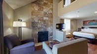 Located at 711 South Main Street in Wichita, Kansas, this two-story suite offers a well-appointed layout designed for comfort and functionality. The main level centers around a striking living area highlighted by soaring vaulted ceilings and a floor-to-ceiling stone-tiled fireplace that serves as a dramatic focal point. Rich hardwood flooring runs throughout, complementing the warm, neutral tones of the space. Comfortable seating surrounds the fireplace, and a flat-screen TV is conveniently positioned nearby. A sliding glass door brings in natural light and provides access to an outdoor area. The fully equipped kitchen on the main level features warm wood cabinetry, stone tile backsplash, granite-style countertops, and black appliances including a range, microwave, dishwasher, and full-size refrigerator — ideal for longer stays. A well-lit vanity area with a large mirror and open shelving connects to a clean, functional bathroom complete with a shower-tub combination and hardwood-style flooring. The upper loft level, accessible via a staircase with wooden railings, houses the sleeping area, which features a queen bed, wall-mounted TV, and windows that allow natural light to filter in. The overall ambiance is cozy yet practical, blending residential warmth with hotel-style convenience.