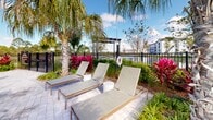 Located at 9500 Grandtree Ave in Venice, Florida, this outdoor amenity space offers a resort-inspired retreat designed for relaxation and leisure. At the heart of the area is a generously sized swimming pool with crystal-clear blue water, framed by a broad paver deck that accommodates rows of modern sling-style lounge chairs ideal for sunbathing. The pool's entry features a gradual shallow zone, making it both accessible and inviting. Surrounding the pool, vibrant tropical landscaping creates a lush, immersive atmosphere, with an abundance of palm trees, red-leafed tropical plants, and dense green foliage that evoke a true Florida resort experience. Paved walkways wind through the greenery, connecting multiple seating and lounging areas, including a shaded pergola structure and intimate outdoor furniture groupings nestled among the palms and colorful plantings. The space is enclosed by a sleek black metal fence, offering privacy and security while maintaining open sightlines to the sky. Multi-story residential buildings frame the backdrop, reinforcing the community setting. Natural light floods the entire area throughout the day, and the combination of lush plantings and open hardscape creates a dynamic, welcoming environment well-suited for outdoor gatherings and quiet relaxation alike.
