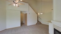 This two-story residence is defined by its open and airy main living area, where vaulted ceilings create a strong sense of vertical space. The room is finished with wall-to-wall neutral carpeting that extends throughout, contributing to a clean and cohesive aesthetic. A ceiling fan with a light fixture is mounted overhead, providing both circulation and ambient lighting, while a pendant light near the staircase adds a decorative accent.

The staircase, featuring wooden handrails, rises along one wall and connects the main level to the upper floor, which is partially visible from below, reinforcing the open feel of the space. A wall cutout or ledge near the upper level suggests a potential pass-through or display area between floors.

Adjacent to the main living area, a hallway leads to additional rooms, with a doorway visible at the end and a floor vent indicating a ducted HVAC system. The walls throughout are finished in a soft, neutral tone, keeping the interior bright and versatile. Overall, the space offers a functional and open layout suited to a variety of living arrangements.