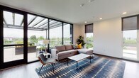 Located at 4018 N Williams Ave in Portland's vibrant Williams Avenue corridor, this contemporary single-floor space offers a sleek, modern design with an open and airy ambiance. The centerpiece of the room is a dramatic floor-to-ceiling black-framed glass wall system that opens directly onto a covered outdoor terrace, flooding the interior with abundant natural light and providing expansive views of the surrounding Portland cityscape and greenery. The smooth white ceiling features recessed lighting, complementing the clean, minimalist aesthetic throughout. Durable wood-style flooring runs across the entire space, grounding the room with warmth and texture. A wall-mounted flat-screen display and a boldly painted dark accent wall provide a clear media and gathering focal point, while a wall-mounted mini-split HVAC unit ensures year-round climate comfort. Multiple entry and exit points, including a marked emergency exit, add to the space's functionality. The interior is accessorized with a bold geometric-patterned area rug in navy and tan tones, a sectional sofa, a marble-top coffee table, and mid-century modern accent seating, creating an inviting and well-appointed lounge environment ideal for communal gatherings or events.