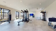 Located at 305 Prine Place in Charlotte, NC, this contemporary fitness center offers a well-appointed, single-level layout designed to support a full range of exercise routines. The space is characterized by its clean, modern aesthetic, featuring light-toned walls, smooth concrete-style flooring, and high ceilings that contribute to an open, airy atmosphere. Abundant natural light floods the interior through large black-framed windows and glass doors, providing views of the surrounding outdoor landscape and greenery.

The facility is equipped with a comprehensive array of professional-grade workout equipment, including a multi-station cable machine, adjustable weight benches, a dumbbell rack with a full range of free weights, medicine balls, kettlebells, and TRX suspension training systems. A dedicated storage rack organizes functional training accessories, keeping the floor space clear and accessible. A heavy bag and resistance rope round out the functional fitness options.

A large wall-mounted flat-screen television serves as a focal point for guided workout programming, while ceiling fans ensure comfortable air circulation throughout. Full-length mirrors along one wall enhance the sense of space and allow users to monitor their form during training. Glass doors open directly to an outdoor area, seamlessly connecting the interior fitness space to the exterior environment.