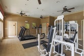 Located at 461 Old Dixie Way in Forest Park, Georgia, this single-floor fitness center is thoughtfully designed to provide a well-rounded workout environment. The space features warm, neutral-toned walls accented by elegant crown molding and classic white baseboards, giving the room a polished, welcoming feel. Multiple ceiling fans are distributed throughout, ensuring consistent airflow and comfort during exercise sessions. The flooring consists of low-pile carpet with protective rubber mats positioned beneath the equipment. A dedicated cardio zone houses multiple treadmills positioned near large windows and glass doors, which flood the area with natural light and offer views of the surrounding outdoor spaces. The strength and multi-use equipment area includes a cable machine, elliptical trainer, weight bench, and free weights, arranged for efficient use of the available floor space. Large framed mirrors line portions of the walls, visually expanding the room and allowing users to monitor their form. Decorative valances in a deep red tone add a touch of warmth and character near the windowed areas. An exit door provides direct access to the exterior. The overall ambiance is bright, functional, and inviting, making this fitness center a well-appointed amenity for its community.