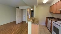 This single-floor residence offers a thoughtfully arranged layout that balances functionality and comfort. The kitchen is outfitted with warm wood cabinetry and tile flooring, and opens to the living area via a peninsula-style breakfast bar topped with granite, providing both a casual dining surface and a natural division between spaces. The living area features rich hardwood flooring that adds warmth and visual continuity throughout the space. A doorway leads to a hallway that connects to additional rooms, including what appears to be a bedroom with a spacious closet visible through the open door. Natural light filters in through a window at the end of the hallway, brightening the interior. The walls are finished in a soft neutral tone, creating a clean and inviting atmosphere throughout. Flush-mount ceiling fixtures provide consistent ambient lighting across the space. The open sightlines between the kitchen, living area, and hallway contribute to a connected, airy feel, making the home feel welcoming and easy to navigate. Overall, the space presents a practical and comfortable layout well-suited for everyday living.