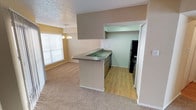 This single-floor residence offers a practical and open layout designed to maximize functionality. The living and dining area flows seamlessly from the kitchen, creating a connected, communal atmosphere. A peninsula-style kitchen counter with a built-in sink and dark laminate countertop serves as the natural dividing point between the kitchen and the carpeted living space. The kitchen area features hardwood-style flooring, dark cabinetry, and black appliances, providing a functional workspace with straightforward access to the adjacent dining and living areas. An accent wall in a warm taupe tone adds a subtle visual contrast to the otherwise neutral color palette throughout the space. The dining area benefits from natural light streaming in through a large window fitted with horizontal blinds, and is complemented by a chandelier overhead, lending a modest decorative touch. The textured popcorn ceiling is consistent throughout, and the soft, neutral carpet in the living area provides a comfortable underfoot feel. A hallway visible from the main space suggests additional rooms beyond, contributing to the overall sense of a well-organized, easy-to-navigate floor plan.