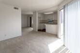 Located at 1256 W Arrow Hwy in Upland, California, this single-floor unit offers a straightforward and functional layout spread across an open, connected living space. The main living area is generously proportioned, featuring light-toned carpeting and a textured ceiling that runs throughout. A sliding glass door allows natural light to pour in while providing access to the outdoors. The kitchen is positioned adjacent to the living area and features white cabinetry with ample upper and lower storage, light speckled countertops, and a full suite of white appliances including an electric range, over-the-range microwave, and dishwasher. A double-basin sink sits beneath a window, adding natural light to the workspace. The kitchen floor is finished in warm hardwood-style flooring, which also extends into the bathroom. The bedroom is spacious and carpeted, offering a sliding-door closet with built-in shelving and an adjacent vanity or dressing area. The bathroom is clean and simply appointed, featuring a full soaking tub with shower, wood-style flooring, and bright white walls. Central HVAC vents are present throughout, ensuring year-round comfort. The unit presents a clean, neutral palette throughout, making it a versatile and move-in ready space.