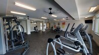 This fitness center offers a spacious, single-floor layout designed to accommodate a wide variety of workout routines. The room features a dark, rubberized flooring ideal for both cardio and strength training activities, providing durability and safety underfoot. Multiple ceiling fans are evenly distributed throughout the space, ensuring consistent airflow and comfort during exercise sessions.

The facility is well-lit with a combination of overhead fluorescent lighting and natural light filtering in through windows near the entrance, creating a bright and energizing atmosphere. A row of elliptical machines and other cardio equipment lines one side of the room, while free weights, benches, and resistance machines are organized toward the back and center of the space.

Wall-mounted mirrors along one section enhance the sense of openness and allow users to monitor their form during workouts. Colorful abstract artwork decorates the walls, adding a touch of visual interest to the otherwise functional environment. The overall ambiance is clean, modern, and practical, making it a well-rounded facility suited for a range of fitness levels and training goals.