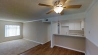 This single-story space showcases an open-concept design that seamlessly connects the living area and kitchen. The living room features a combination of wood-look hardwood flooring and carpeted sections, creating distinct yet flowing zones within the shared space. A ceiling fan with a light fixture is centrally mounted on the textured popcorn ceiling, providing both ventilation and ambient lighting. Crown molding adds a classic finishing detail along the ceiling perimeter.

The kitchen is positioned adjacent to the living area and is accessible via a raised breakfast bar counter with a gray surface, ideal for casual dining or additional workspace. White cabinetry lines the kitchen walls, offering ample storage, while a pendant light hangs above, adding focused illumination. The kitchen also features a sink visible from the pass-through area.

Neutral-toned walls throughout the space contribute to a clean, versatile aesthetic. A structural column near the kitchen transition adds an architectural detail that subtly defines the two areas. The overall design is practical and straightforward, offering a functional layout suited to everyday living.