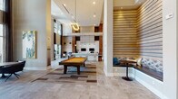 This single-floor community clubhouse in Dayton, Kentucky, is designed with a contemporary aesthetic that blends warm wood accents, neutral stone-tiled flooring, and dramatic double-height ceilings throughout. The space is open and expansive, with distinct zones that serve a variety of social and recreational purposes.

Upon entry, French doors with glass panels open into a bright foyer finished in soft blue-gray tones, accented by horizontal shiplap wall detailing and a built-in upholstered bench nook. The main lounge area features two generously sized sofas flanking a central coffee table beneath a statement circular chandelier. A wall-mounted television is framed by a clean white surround, with horizontal slatted wood panels extending floor to ceiling on either side, adding texture and warmth.

Adjacent to the lounge, a full billiards table anchors the game area, complemented by a bar-style counter with barstool seating and pendant lighting overhead. Dual monitors and workstation seating nearby provide a functional business center space. Floor-to-ceiling windows at the far end of the room flood the interior with natural light and offer unobstructed outdoor views, while framed artwork enhances the gallery-like wall treatments throughout.