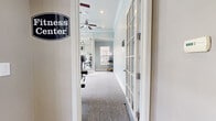 This fitness center is a thoughtfully designed workout space that combines functionality with a welcoming aesthetic. The room is finished with soft, light gray carpeting underfoot and walls painted in a cool, calming blue-gray tone, accented by crisp white crown molding and baseboards that lend a polished, refined look throughout.

The space is well-lit by a combination of recessed ceiling lights and multiple large windows, including a prominent arched window, which allow abundant natural light to pour in and provide views of the surrounding outdoor environment. Ceiling fans positioned throughout the room ensure consistent airflow during workouts.

A full wall of floor-to-ceiling mirrors visually expands the space and serves as a practical training aid. The fitness center is equipped with a variety of cardio machines, including treadmills, a stationary upright bike, and a cycling bike, as well as multi-station cable and weight machines designed to support comprehensive strength training. Wall-mounted flat-screen televisions are positioned for easy viewing during exercise sessions.

Entry to the fitness center is through elegant French-style glass-paned doors, clearly marked with a "Fitness Center" sign, creating a clean and professional transition from the adjacent corridor with its tile flooring. The overall ambiance is bright, airy, and purpose-built to support an active and healthy lifestyle.
