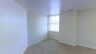This single-floor space presents a clean, minimalist aesthetic defined by bright white walls and neutral carpet flooring that runs throughout. The room pictured showcases a classic double-hung window that allows natural light to fill the interior, creating a warm and airy atmosphere. The white painted walls and ceiling contribute to a sense of openness, while the light-toned carpet provides a soft, comfortable underfoot experience. Ceiling-mounted lighting fixtures and standard HVAC vents are integrated seamlessly into the design, ensuring functional comfort throughout the space. The clean baseboard trim running along the perimeter of the room adds a subtle architectural finish. The overall ambiance is straightforward and practical, offering a neutral canvas that can easily accommodate a variety of uses or personal styles. The absence of clutter and the uniform color palette make the space feel open, tidy, and move-in ready. Whether used as a living area, bedroom, or multi-purpose room, this space provides a solid, well-maintained foundation with ample natural light and a calm, understated character.
