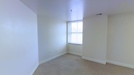 This single-floor space presents a clean, minimalist aesthetic defined by bright white walls and neutral carpet flooring that runs throughout. The room pictured showcases a classic double-hung window that allows natural light to fill the interior, creating a warm and airy atmosphere. The white painted walls and ceiling contribute to a sense of openness, while the light-toned carpet provides a soft, comfortable underfoot experience. Ceiling-mounted lighting fixtures and standard HVAC vents are integrated seamlessly into the design, ensuring functional comfort throughout the space. The clean baseboard trim running along the perimeter of the room adds a subtle architectural finish. The overall ambiance is straightforward and practical, offering a neutral canvas that can easily accommodate a variety of uses or personal styles. The absence of clutter and the uniform color palette make the space feel open, tidy, and move-in ready. Whether used as a living area, bedroom, or multi-purpose room, this space provides a solid, well-maintained foundation with ample natural light and a calm, understated character.