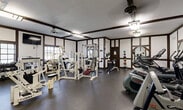 Located at 3710 Inverrary Dr in Lauderhill, Florida, this fitness center offers a well-appointed workout facility designed to accommodate a wide range of exercise needs. The space features a distinctive Tudor-style architectural design, with dark wood beam accents contrasting against white walls, lending the gym a classic and structured aesthetic that sets it apart from typical fitness facilities.

The gym is outfitted with a comprehensive array of equipment, including multiple Cybex strength machines, cable and pulley systems, a multi-station weight machine, and a selection of free weights and dumbbells along one wall. Cardio options are equally well-represented, with treadmills, elliptical trainers, and stationary bikes positioned throughout the space. Medicine balls and a weight bench round out the strength training area.

Large mirrors are strategically placed along the walls, enhancing the sense of openness and allowing users to monitor their form during workouts. Natural light filters in through windows, supplemented by overhead fluorescent and pendant-style lighting fixtures. A wall-mounted flat-screen TV provides entertainment during workouts. The durable rubber flooring throughout ensures safety and comfort. The space is efficiently organized to maximize functionality, making it a practical and fully equipped fitness resource.