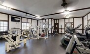 Located at 3710 Inverrary Dr in Lauderhill, Florida, this fitness center offers a well-appointed workout facility designed to accommodate a wide range of exercise needs. The space features a distinctive Tudor-style architectural design, with dark wood beam accents contrasting against white walls, lending the gym a classic and structured aesthetic that sets it apart from typical fitness facilities.

The gym is outfitted with a comprehensive array of equipment, including multiple Cybex strength machines, cable and pulley systems, a multi-station weight machine, and a selection of free weights and dumbbells along one wall. Cardio options are equally well-represented, with treadmills, elliptical trainers, and stationary bikes positioned throughout the space. Medicine balls and a weight bench round out the strength training area.

Large mirrors are strategically placed along the walls, enhancing the sense of openness and allowing users to monitor their form during workouts. Natural light filters in through windows, supplemented by overhead fluorescent and pendant-style lighting fixtures. A wall-mounted flat-screen TV provides entertainment during workouts. The durable rubber flooring throughout ensures safety and comfort. The space is efficiently organized to maximize functionality, making it a practical and fully equipped fitness resource.