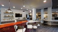 Located at 4625 Fairview Dr in Wilmington, NC, this single-floor commercial space offers a sleek, modern aesthetic designed to impress. The open-concept layout seamlessly connects multiple functional zones, creating a dynamic and inviting environment throughout.

A standout feature is the bold bar-height counter with a vibrant orange top, accented by a striking red brick base and lined with contemporary metal and upholstered bar stools. Overhead, a mix of pendant lighting — including an eye-catching chrome globe chandelier and an industrial-style dome pendant — adds character and warmth to the space.

The rear counter area is equipped with built-in cabinetry in a dark finish, a wall-mounted flat-screen television, and what appears to be refreshment or service station amenities, suggesting a hospitality or client-facing function. White brick accent walls provide a textural contrast against the smooth grey ceiling and tiled flooring, lending the space an urban, refined feel.

Beyond the main counter, the open floor plan extends into additional seating and workspace areas, with ample room for collaborative or individual use. Recessed lighting throughout ensures the entire space is well-lit, while the cohesive modern design palette of grey, white, and bold accent colors ties every area together into a polished, professional environment.
