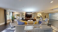 Located at 124 Ellington Blvd in Gaithersburg, Maryland, this thoughtfully designed single-floor amenity space offers a sophisticated and inviting environment for social gatherings and relaxation. The open-concept layout seamlessly connects multiple functional zones, creating a fluid and spacious feel throughout.

The main lounge area is elegantly appointed with contemporary furnishings, including plush neutral sofas accented with mustard and cobalt blue throw pillows, arranged around a geometric patterned area rug. A dark wood media wall with integrated shelving and a large flat-screen TV serves as a refined focal point, flanked by curated decorative art and sculptural accents. Statement pendant and drum lighting fixtures illuminate the space warmly, while large French doors and oversized windows invite abundant natural light and views of the surrounding greenery.

The bar and kitchen area showcases a curved, white-topped island with sleek bar seating, dark espresso cabinetry, and a striking mosaic tile backsplash. Pendant lights drop from an architecturally detailed tray ceiling, enhancing the upscale ambiance. Stainless steel appliances complete the fully functional setup. Dark hardwood-style flooring flows cohesively throughout the entire space. A billiards table rounds out the entertainment offerings, making this versatile venue ideal for community events and casual socializing alike.