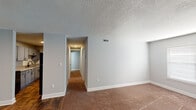 This single-story unit in South Carolina presents a thoughtfully updated interior with a cohesive, neutral color palette of soft gray walls and crisp white trim throughout. The main living area features a combination of rich, dark wood-look flooring and wall-to-wall carpeting, with a textured ceiling overhead and a front entry door flanked by a window that allows natural light into the space. The layout transitions smoothly from the living area into the kitchen, which is equipped with gray shaker-style cabinetry accented by matte black hardware, granite-look countertops, a white subway tile backsplash, and black appliances including an electric range. A hallway extends from the living space toward the private rooms, lined with carpeting and leading to a walk-in closet outfitted with dual shelving and hanging rods on opposing walls, providing ample storage. The bathroom features wood-look tile flooring, a gray vanity with a granite countertop, and a clean white tub-shower surround with chrome fixtures. The unit's consistent finishes and practical layout make it a functional and well-appointed living space.