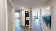 This thoughtfully designed single-floor residence offers a comfortable and functional layout with two bedrooms, a full bathroom, a modern kitchen, and a bright living area. Wood-style flooring flows through the main hallway and living spaces, while plush carpet adds warmth to the bedrooms.

The kitchen is outfitted with crisp white shaker cabinetry, subway tile backsplash, stone countertops, stainless steel appliances, and a built-in wine rack, creating a polished and practical space for cooking and entertaining. The living room is bathed in natural light from large windows and a glass door that opens to a private balcony, offering views of the surrounding community.

The two bedrooms provide comfortable retreats, each featuring carpeted flooring and ample space for furnishings. The full bathroom includes a clean white vanity with stone countertop, a large mirror, herringbone-patterned tile flooring, and a tub-shower combination.

A central hallway connects all areas of the residence and features a dedicated in-unit laundry closet housing a full-size stacked washer and dryer, conveniently tucked behind bi-fold louvered doors. Recessed lighting and neutral wall tones throughout contribute to a cohesive, modern aesthetic.