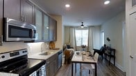 This well-appointed single-floor residence offers a seamlessly integrated open-concept layout that connects the kitchen, dining, and living areas into one cohesive and functional space. The kitchen is outfitted with sleek gray cabinetry featuring frosted glass upper panels, light stone countertops, a subway tile backsplash, and stainless steel appliances including a built-in microwave and dishwasher. Under-cabinet lighting adds a warm, practical glow to the workspace, which centers around a stainless steel sink. A dining area sits adjacent to the kitchen, furnished with a modern bar-height table and seating that transitions naturally into the open living space beyond. Rich hardwood-style flooring runs throughout, grounding the contemporary aesthetic. The living area benefits from generous natural light streaming through multiple windows dressed with white curtains and blinds, creating a bright and airy atmosphere. A ceiling fan with an integrated light fixture provides both comfort and additional illumination. The overall ambiance is clean, modern, and inviting, with a neutral color palette accented by contemporary furnishings and thoughtful design details that make the space both practical and visually appealing.