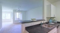 This single-floor unit showcases an open-concept design that seamlessly connects the kitchen and living areas into one cohesive space. The kitchen features dark countertops that contrast with the light, neutral walls, and includes a peninsula with an overhang that serves as both a workspace and a casual dining or serving surface. A stainless steel sink is integrated into the countertop, adding a functional and clean aesthetic. The flooring transitions from a hard surface in the kitchen area to soft carpeting in the living space, creating distinct yet connected zones. The living area is bathed in natural light streaming through a large window, which also offers a view of the surrounding exterior. A ceiling fan is centrally mounted above the living area, providing both air circulation and a subtle design element. The walls are finished in a soft, neutral tone throughout, contributing to a clean and airy atmosphere. Electrical outlets are conveniently distributed along the walls. The overall layout is practical and efficient, with an open flow that maximizes the sense of space within the unit.